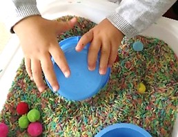 criança manuseia arroz e bolinhas de feltro coloridas dentro de um alguidar.